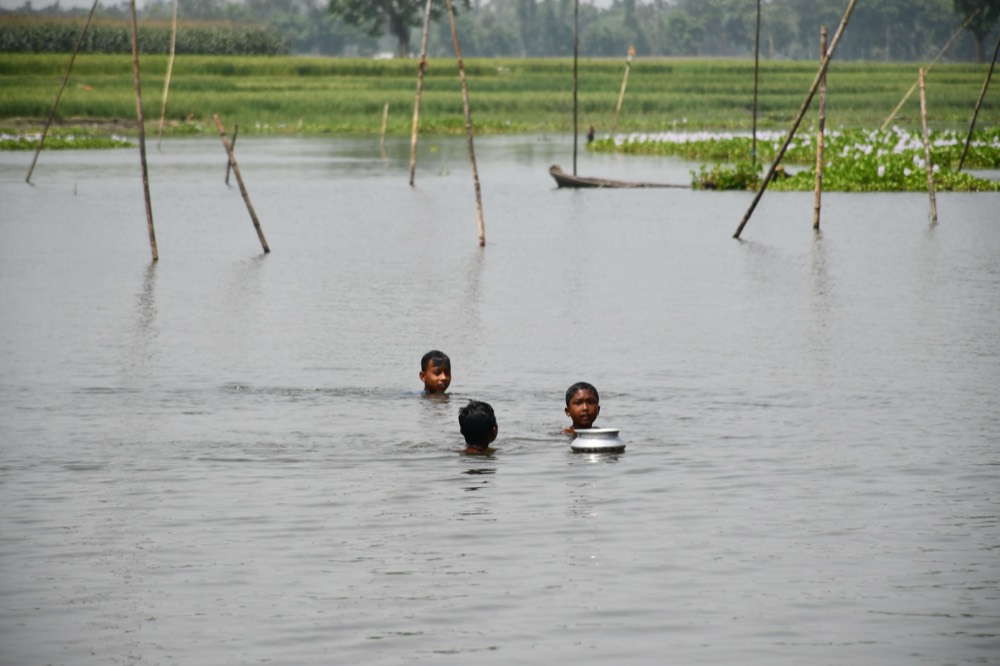 Children wade through chest-deep floodwater carrying a cooking pot across submerged fields